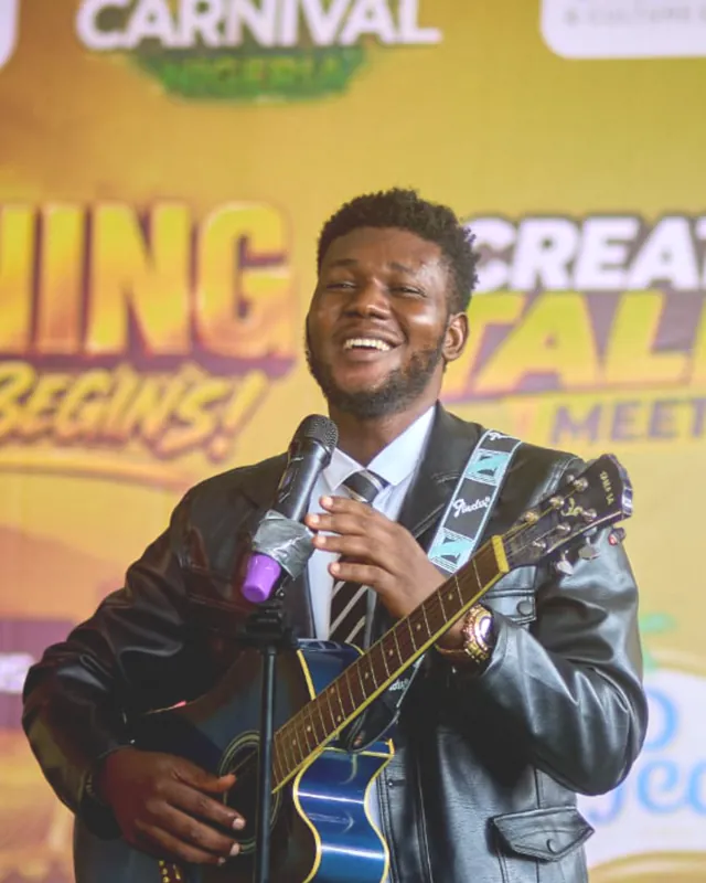 A young musician singing with his guitar on stage at The Awakening, the carnival backdrop behind him